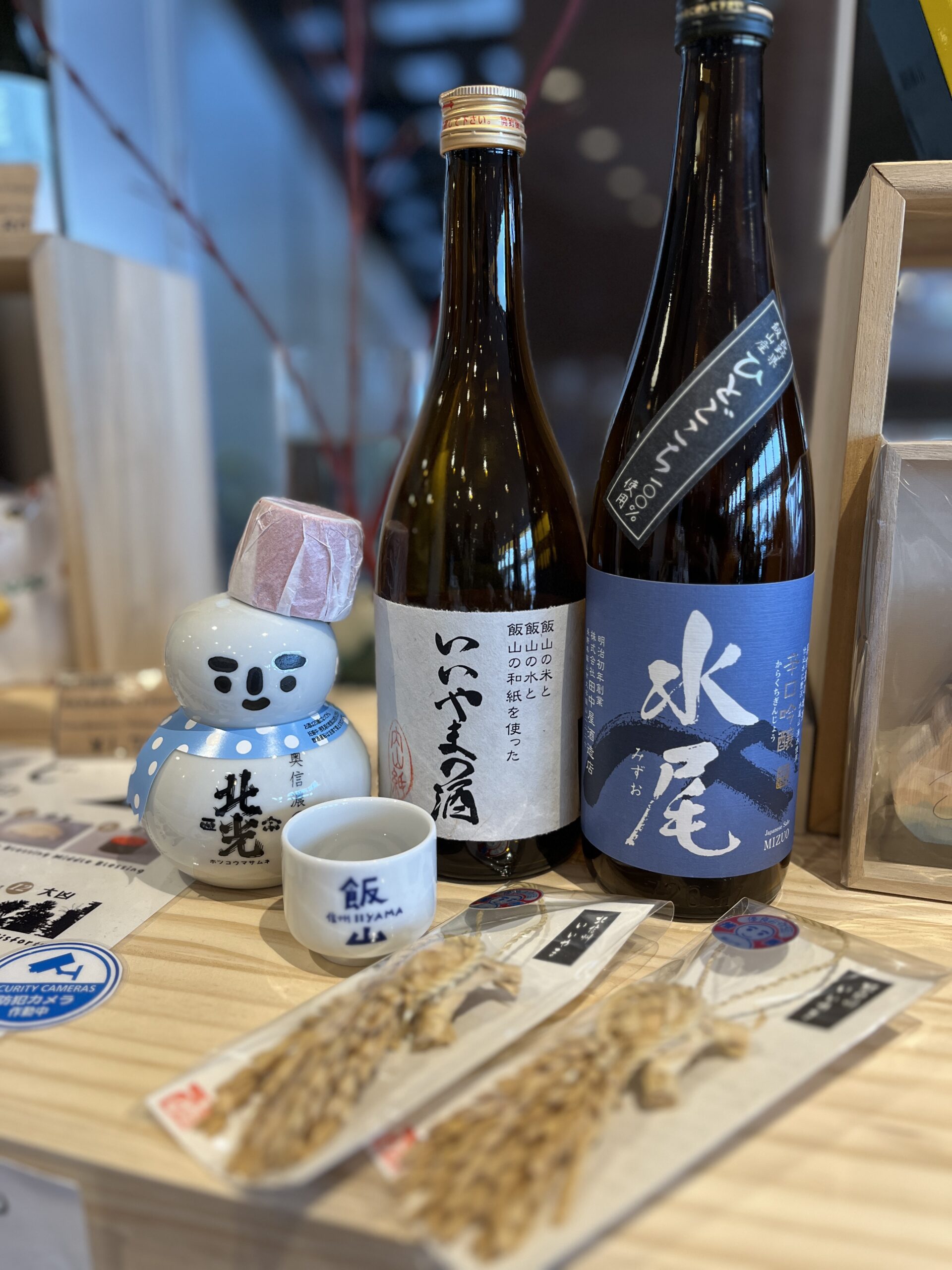 Assorted bottles of local Japanese sake displayed on a wooden counter alongside small souvenir items, including a snowman-shaped sake bottle and packaged snacks.