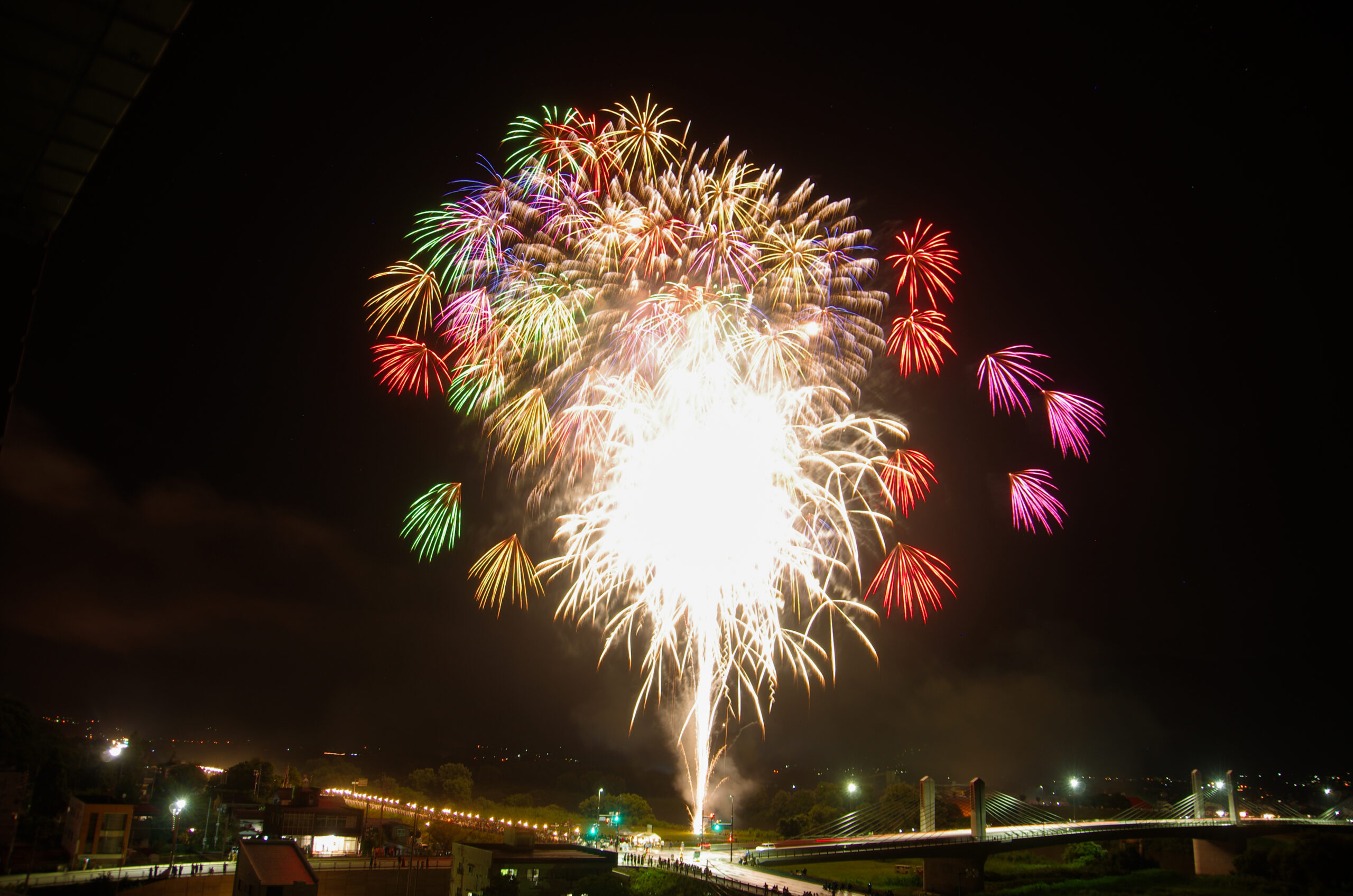 Colorful fireworks bursting over the Chikuma River with a bridge and night skyline in the background